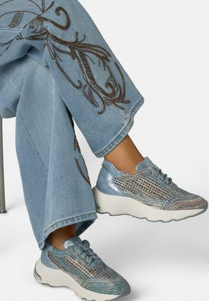 Jambes croisées portant un jean bleu clair avec une broderie marron et des baskets bleues métalliques avec des semelles blanches et des détails en mesh.
