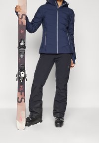 Personne portant une veste de ski bleu marine, un pantalon de ski noir et des chaussures de ski tenant un ski rose et noir verticalement sur un fond uni.