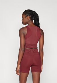 Haut de sport bordeaux avec un panneau en mesh et un banderole de marque ; associé à un short assorti. Son design ajusté met en valeur les contours du corps.