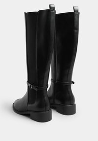 Svarta knähöga stövlar med en slät läderfront och texturerade tygsidor, med en spänndetalj och en låg, tjock klack.