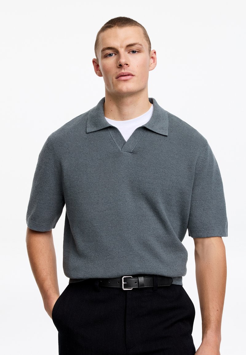 Homme portant un polo gris en maille à manches courtes sur un T-shirt blanc, pantalon noir et ceinture noire, debout avec une main dans la poche.