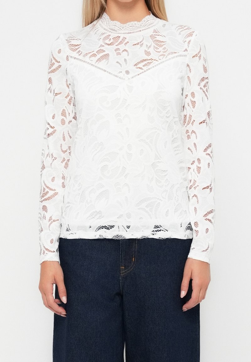 Blouse en dentelle blanche à manches longues, motif floral, col montant, et bords festonnés ; associée à un pantalon large en denim foncé.