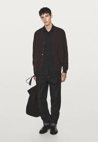 Brauner Cardigan über einem schwarzen Button-Up-Shirt, kombiniert mit dunklen Jeans und schwarzen Schuhen. Hält eine braune Tragetasche und zeigt einen entspannten Look.