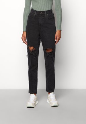 Personne portant un jean noir taille haute déchiré avec des trous aux genoux, associé à un haut vert côtelé à manches longues et des baskets blanches, debout.