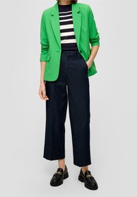 Person klädd i grön kavaj över randig skjorta, mörka vida capribyxor och svarta loafers med gulddetaljer.