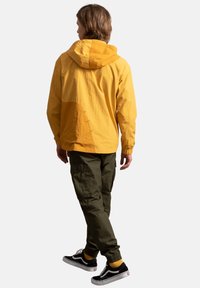 Geel gewatteerd jack met textuur en een oversized pasvorm, gecombineerd met olijfgroene cargo-broeken en zwarte sneakers. Accenten zijn onder andere zijzakken.