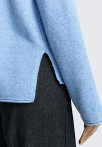 Helles blaues Strickpullover mit strukturiertem Finish, mit einer High-Low-Saum und einem seitlichen Schlitz, kombiniert mit dunklen Denim-Hosen.