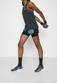 Svart idrottstopp med tre ränder logotyp, gröna och svarta mönstrade shorts, svarta strumpor och grå löparskor med blå detaljer.