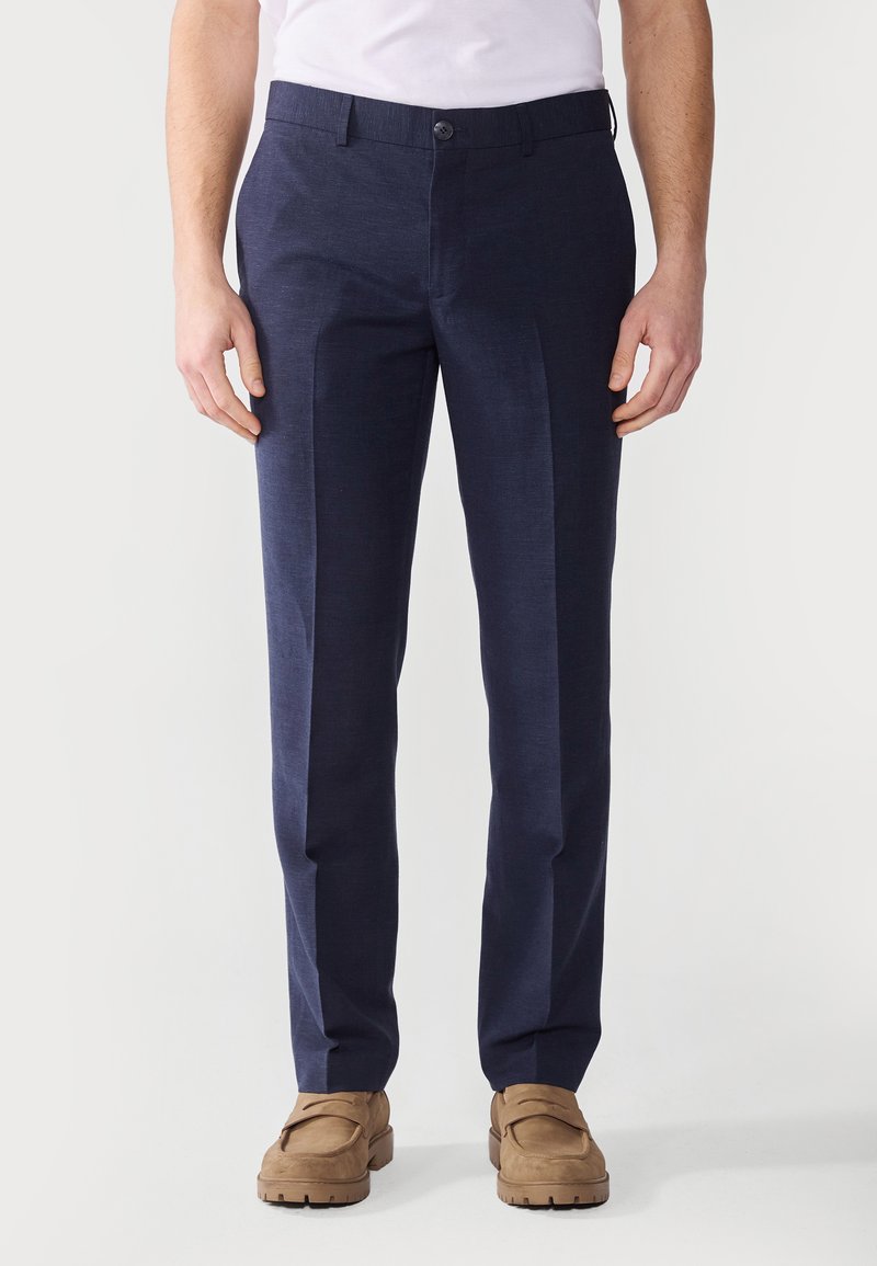 Pantalons taillés bleu marine en tissu lisse, avec une taille plate, des passants de ceinture et une fermeture à un bouton, accompagnés de chaussures brunes.