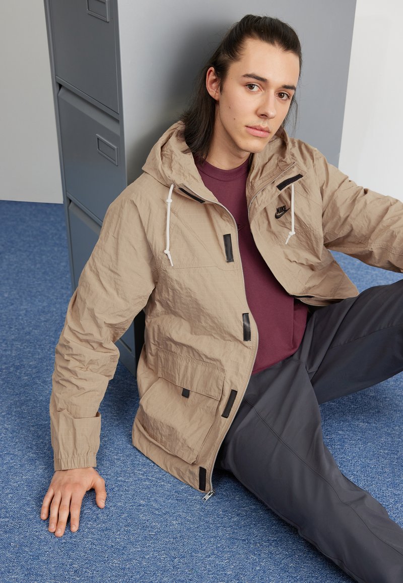 Chaqueta ligera beige con capucha, bolsillos frontales y acentos negros. Llevada sobre una camisa marrón y combinada con pantalones oscuros.