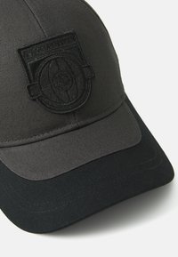 Svart baseballcap med teksturert overflate, buet skjerm og en preget logo-lapp foran. Flat, kontrastfarget svart visir.