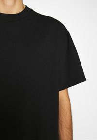 T-shirt à col rond et manches courtes porté par une personne, montrant le haut du torse et une partie du bras droit sur fond blanc.