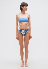 Γυναίκα που στέκεται ξυπόλητη με μπλε μπικίνι με σχέδιο στην κορυφή και μπλε φλοράλ ψηλοκομμένα κάτω, κοιτάζοντας προς τα δεξιά της, με λευκό απλό φόντο.