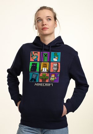 Granatowa bluza z kapturem z pikselowym wzorem postaci z gry Minecraft, wyświetlana w siatce, z logo na dole. Wykonana z bawełny.