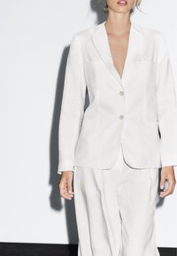 Donna che indossa un blazer bianco su misura con rever e pantaloni bianchi larghi abbinati, in piedi contro uno sfondo chiaro e semplice.