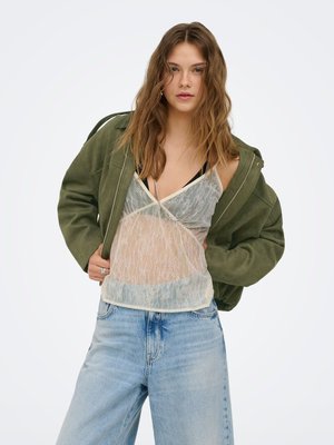Jeune femme aux cheveux bruns lâchés, portant une veste verte sur un top en dentelle transparente et un jean taille haute bleu clair, posant sur fond blanc.
