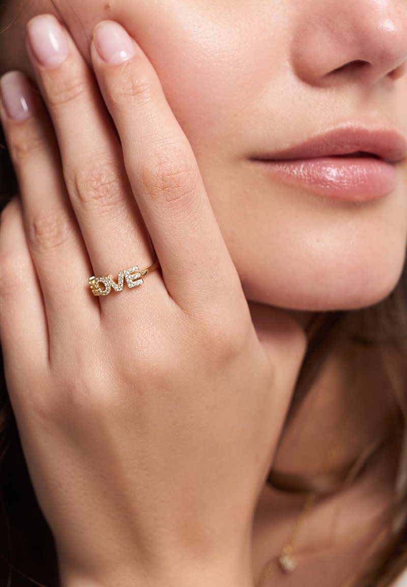 Goldring mit dem Wort "LIEBE" in Großbuchstaben, verziert mit kleinen klaren Steinen, getragen an einer Hand mit hellen Nägeln.