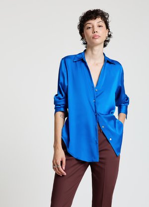 Blue satin blouse with a relaxed fit, long sleeves rolled up, pointed collar, and silver buttons. Worn with brown tailored pants.