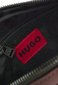 Interior de una bolsa negra con cremallera que muestra una etiqueta roja con "HUGO" en texto negro, con textura suave y detalle de costuras visibles.