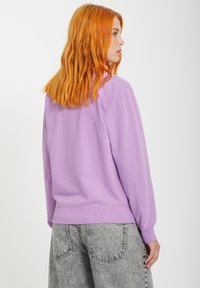 Fioletowa bluza z długimi rękawami, prążkowanymi mankietami i pasem. Noszona z jasnoszarymi, fakturowanymi spodniami o wysokim stanie. Prosty design.