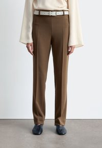 Brown tailored trousers with a smooth texture, featuring a white belt. Worn with a cream top and paired with dark blue shoes.