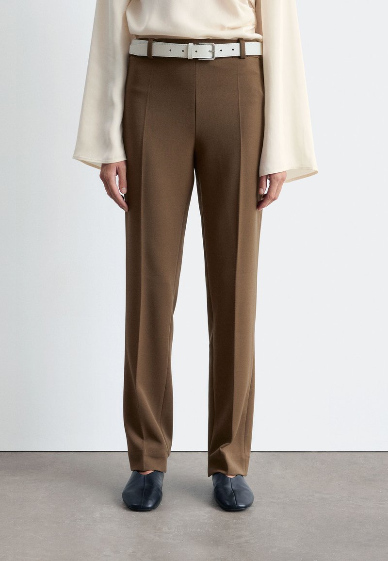 Brown tailored trousers with a smooth texture, featuring a white belt. Worn with a cream top and paired with dark blue shoes.