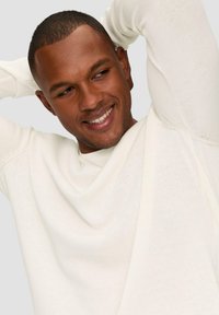 Lichtgekleurde trui met een ontspannen pasvorm, voorzien van een gedrapeerde halslijn en een textuurgebreid patroon, gedragen door een lachende persoon.