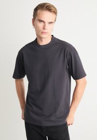 Camiseta de algodón gris oscuro con cuello redondo y mangas cortas. Presenta detalles de costura y un pequeño logo en relieve en el cuello.