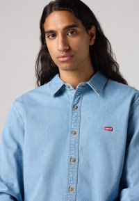 Camicia di jeans azzurro chiaro con bottoni, caratterizzata da cuciture visibili, colletto a punta e un piccolo logo rosso Levi's sul lato sinistro del petto.