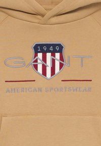 Kohvatu kapuutsiga sviiter, millel on brodeeritud kilbi logo; tekst "GANT 1949" ja "AMERICAN SPORTSWEAR" kontrastsetes värvides.