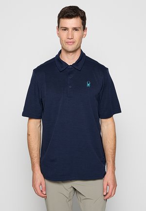 Homme portant un polo bleu marine à manches courtes avec un petit logo araignée turquoise sur le côté gauche de la poitrine.