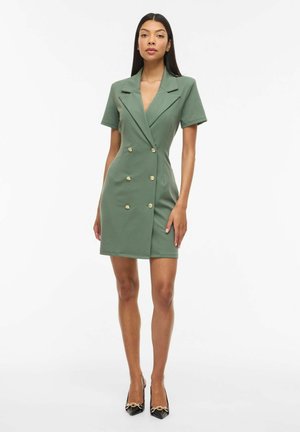 Žena v krátkom outfite na zapínanie s dvoma radmi gombíkov, olivovo zelené s zlatými gombíkmi a čierne špicaté lodičky so zlatým reťazovým detailom.