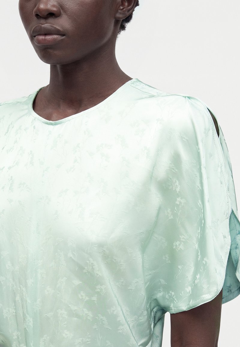 Gros plan d'une personne portant un chemisier en satin vert menthe clair avec des motifs floraux subtils et des manches fendues ouvertes.