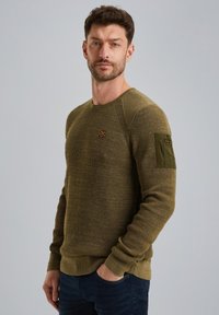 Sudadera verde texturizada con cuello redondo, puños acanalados y un bolsillo tipo parche en la manga. Presenta un pequeño logo en el frente.