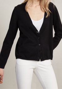 Cardigan noir en tricot avec un col en V, six boutons sur le devant et un ourlet côtelé. Porté par-dessus un débardeur blanc et associé à un jean blanc.