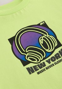Lime zelené tričko s grafickým designem, na kterém jsou fialová sluchátka a text "NEW YORK MUSIC NEVER SLEEPS" ve tučném černém písmu.