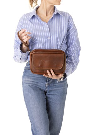 Vrouw in een blauw gestreept shirt en spijkerbroek die een bruine leren polstasje vasthoudt voor een witte achtergrond.