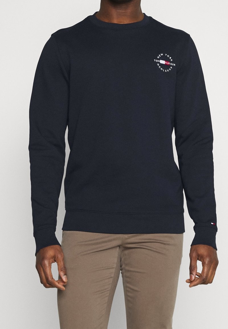 Homme portant un sweat-shirt à manches longues bleu marine avec un logo en texte blanc et rouge sur la poitrine et un pantalon kaki, debout devant un fond uni.