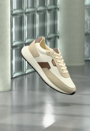 Beżowo-białe sneakersy z zamszu i skóry z brązowymi akcentami i czarną podeszwą, zawieszone nad refleksyjną podłogą blisko szklanych bloków.