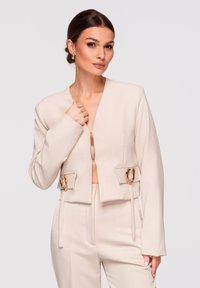 Femme aux cheveux bruns en chignon bas, portant un tailleur crème avec des boucles d’oreilles dorées, une veste courte et un pantalon taille haute sur fond clair.