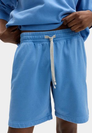 Mand justerer linningen på blå elastikshorts med hvid snøre, iført matchende blå sweatshirt, beskåret ved taljen.