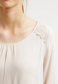 Lysbeige bluse med en rundudskæring, tekstureret stof og delikate blondedetaljer på skulderen. Blødt, flydende materiale.