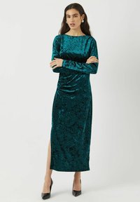 Tyrkysové sametové šaty s dlouhými rukávy, s lodičkovým výstřihem, bočním rozparkem a jemným tečkovaným vzorem. V kombinaci s černými vysokými podpatky.