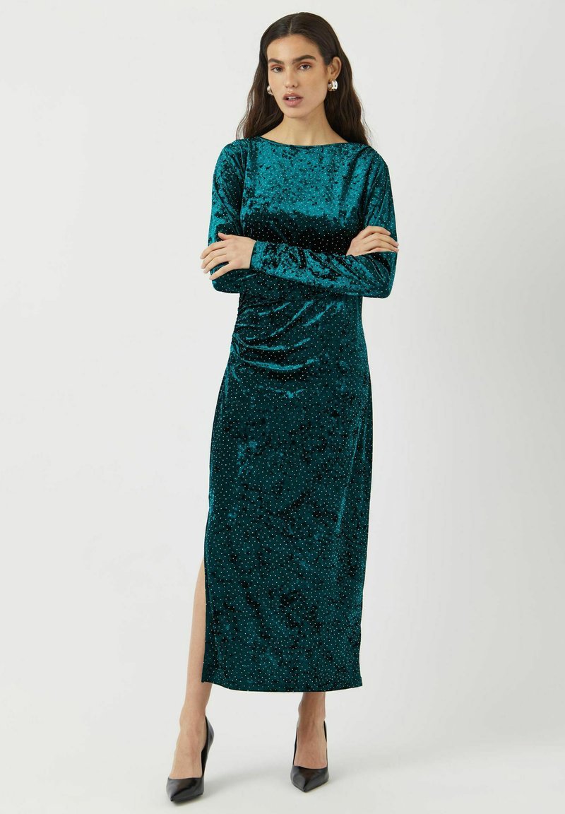 Tyrkysové sametové šaty s dlouhými rukávy, s lodičkovým výstřihem, bočním rozparkem a jemným tečkovaným vzorem. V kombinaci s černými vysokými podpatky.