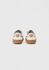 Zapatillas blancas y lila claro con suela de goma, que presentan un logo dorado en el talón y el lateral, material de ante y tela con contraste de texturas.