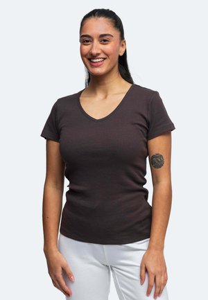 Giovane donna con capelli scuri raccolti, sorridente, che indossa una maglietta a scollo a V scura e pantaloni chiari, mostrando un tatuaggio sul braccio sinistro superiore.