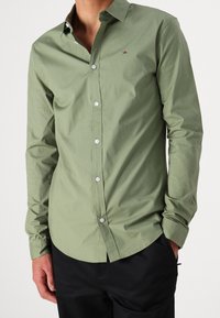 Camicia button-up a maniche lunghe in cotone verde oliva, con colletto classico, bottoni bianchi e un piccolo logo sul petto.