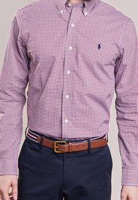 Röd och marinrutigt skjorta med knappad krage, långa ärmar och en liten logotyp, matchad med marinblå byxor och ett brunt bälte.