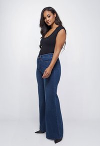 Zwarte bodysuit met ronde hals gecombineerd met hoge taille, wijde donkere spijkerbroeken. De spijkerbroeken hebben voorzakken en subtiele stikdetails.