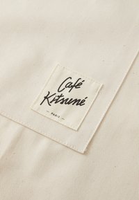 Beige stoff med en subtil stripete tekstur, som har en sydd firkantet etikett som leser "Café Kitsuné Paris" i svart skrift.